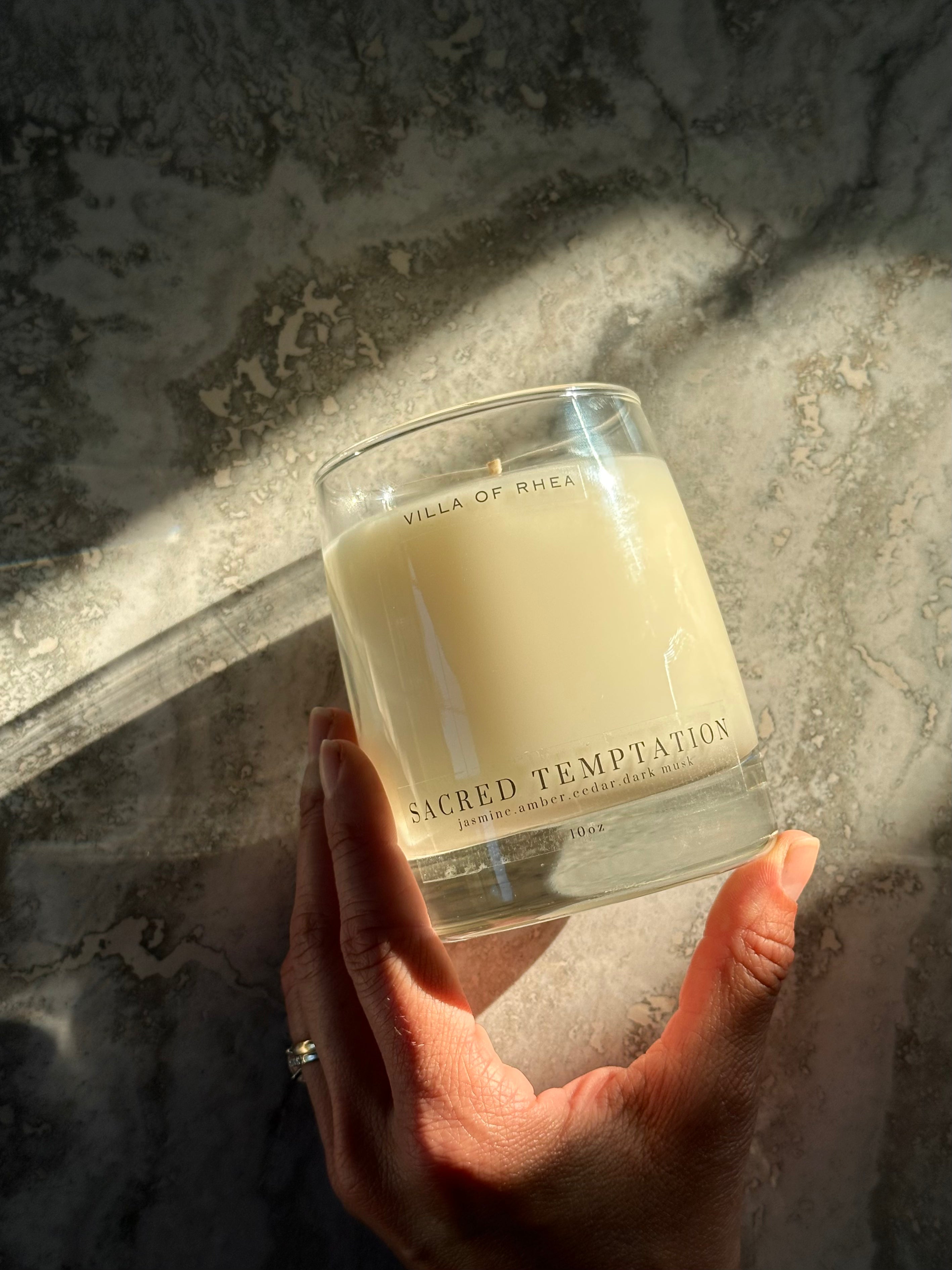 Hand holding a 'Sacred Temptation' candle against a textured wall. Whiskey glass candle. jasmine, amber, cedar, dark musk scent. Vintage style. greek mythology inspired. luxurious soy wax candies. renaissance art. 