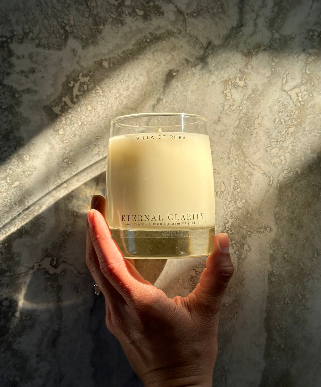 Hand holding a 'Eternal Clarity' candle against a textured wall. eucalyptus, lemon, spearmint and juniper scent. Vintage style. greek mythology inspired. luxurious soy wax candies. renaissance art. 