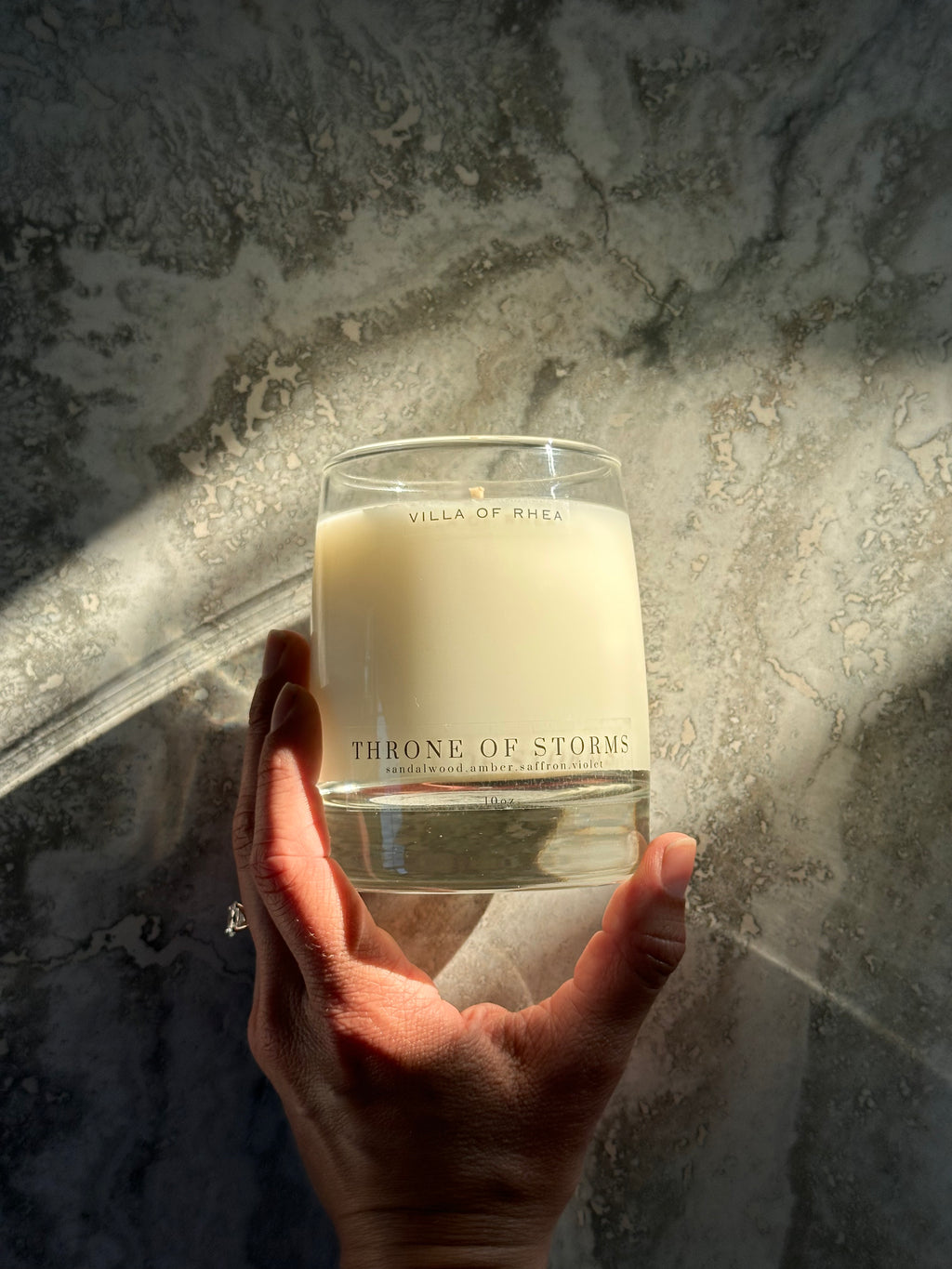 Hand holding a 'Throne of Storms' candle against a textured wall. Whiskey glass candle. sandalwood, amber, saffron and violet scent. Vintage style. greek mythology inspired. luxurious soy wax candies. renaissance art. 