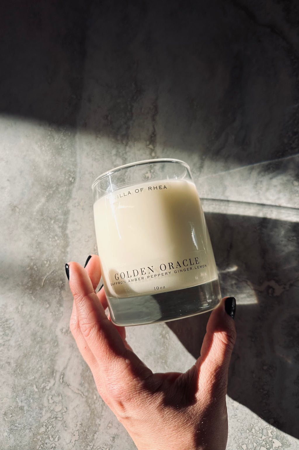 Hand holding a 'Golden Oracle' candle against a textured wall. Whiskey glass candle. Saffron, amber, peppery ginger and lemon scent. Vintage style. greek mythology inspired. luxurious soy wax candies. renaissance art. 