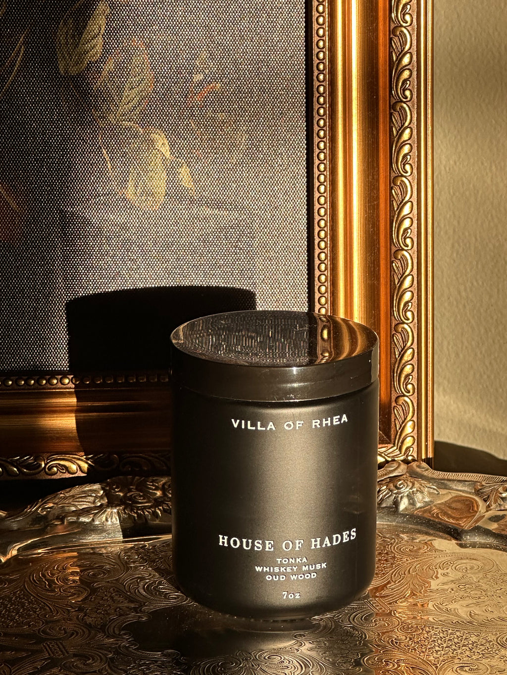 black candle House of Hades candle on a decorative surface with a mirror in the background. tonka, whiskey musk and oud wood scent.Vintage style. greek mythology inspired. luxurious soy wax candies.