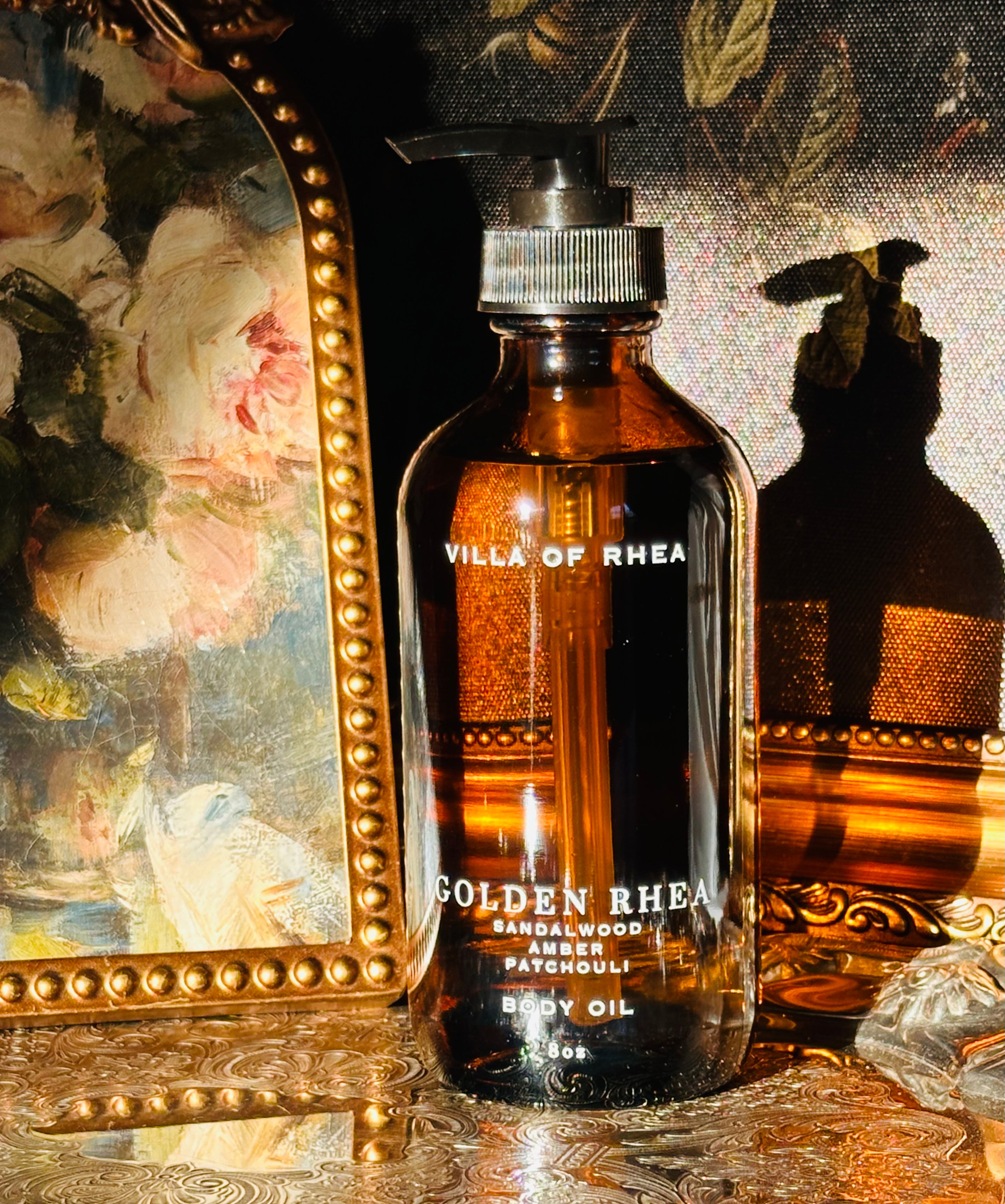 Bottle of  Golden Rhea Amber and Patchouli Scented body oil on a decorative tray with floral elements and a classical statue in the background. Golden Rhea Body Oil. Sandalwood, Amber & Patchouli Scent. Vintage style. Greek Mythology Inspired. Luxury Body Oils