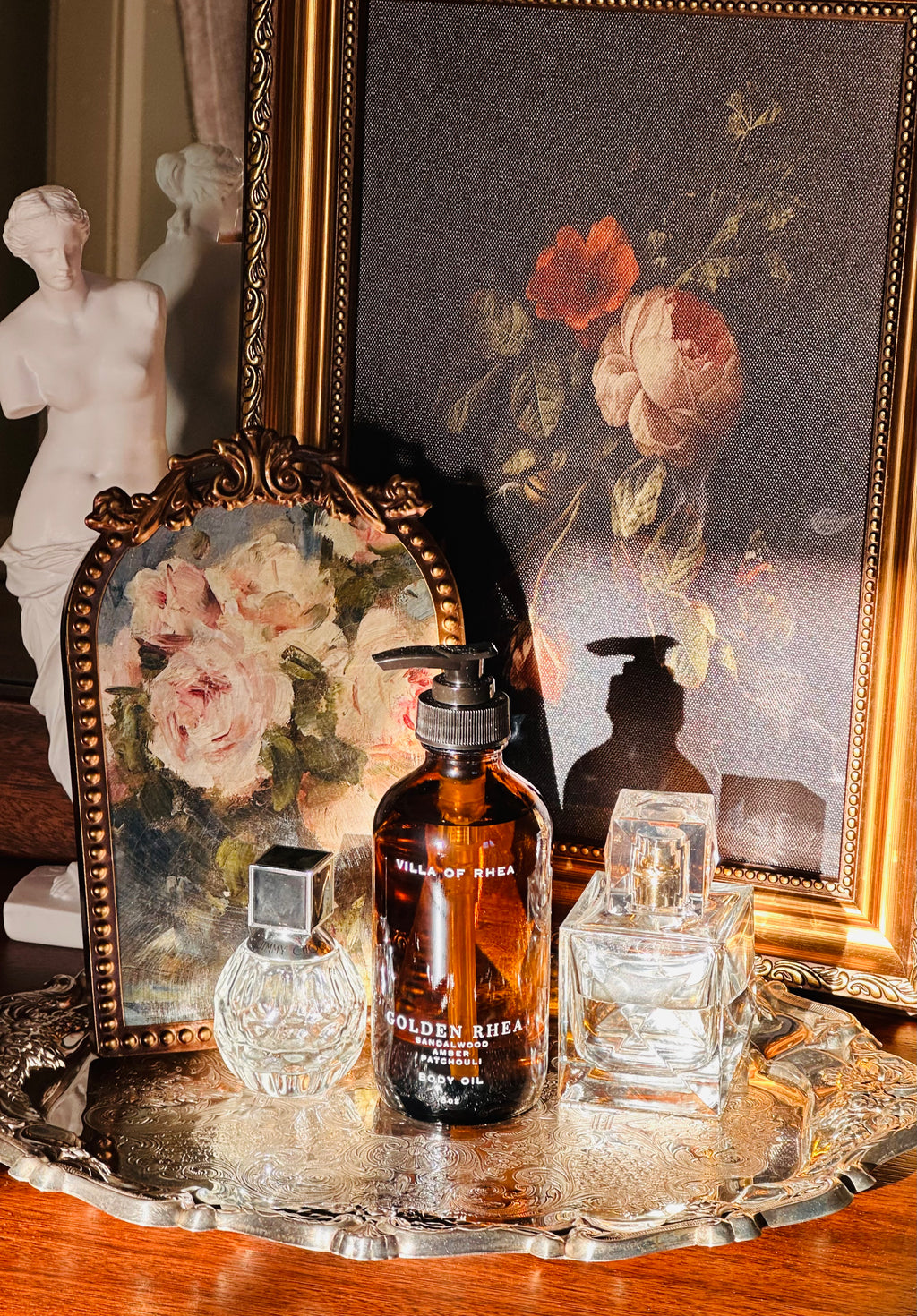 Bottle of  Golden Rhea Amber and Patchouli Scented body oil on a decorative tray with floral elements and a classical statue in the background. Golden Rhea Body Oil. Sandalwood, Amber & Patchouli Scent. Vintage style. Greek Mythology Inspired. Luxury Body Oils