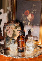 Bottle of  Golden Rhea Amber and Patchouli Scented body oil on a decorative tray with floral elements and a classical statue in the background. Golden Rhea Body Oil. Sandalwood, Amber & Patchouli Scent. Vintage style. Greek Mythology Inspired. Luxury Body Oils
