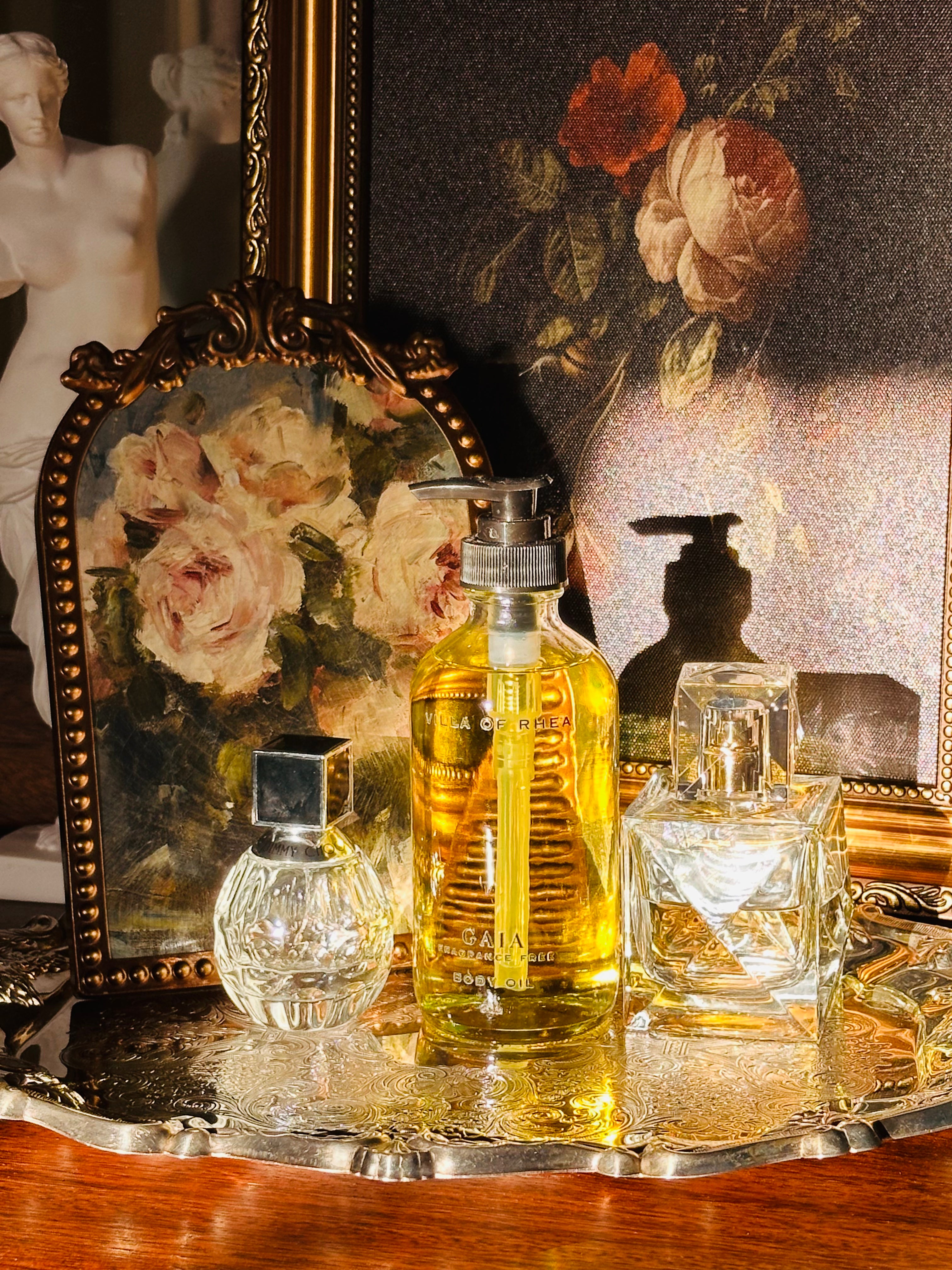 Collection of perfume bottles on a decorative tray with a classical painting in the background. Gaia fragrance free body oil.  on a decorative tray with floral elements and a classical statue in the background. Vintage style. Greek Mythology Inspired. Luxury Body Oils