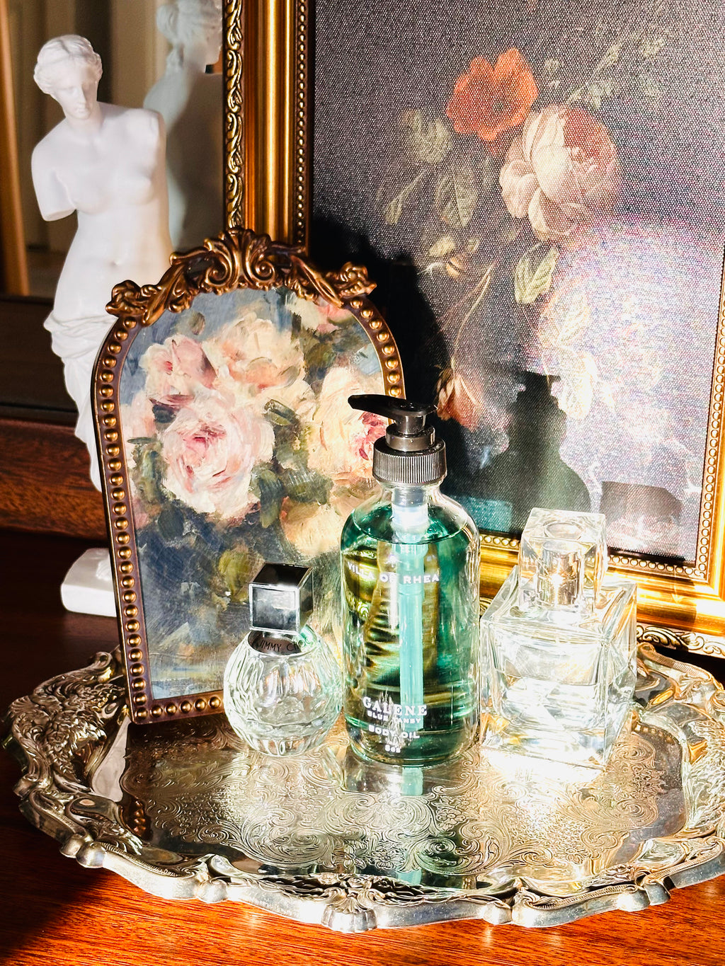 Collection of perfume bottles on a decorative tray with a classical painting in the background. Galene blue tansy body oil.  on a decorative tray with floral elements and a classical statue in the background. Vintage style. Greek Mythology Inspired. Luxury Body Oils