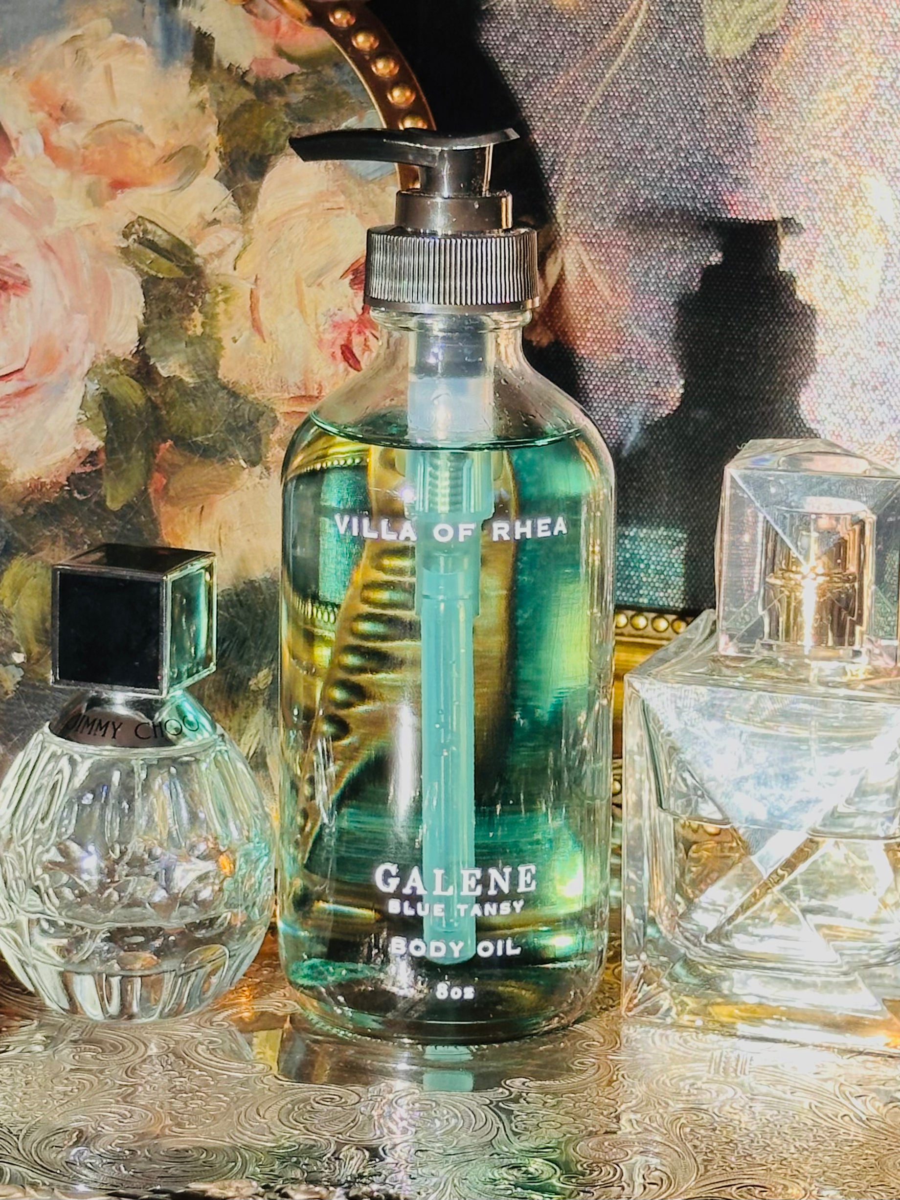Collection of perfume bottles on a decorative tray with a classical painting in the background. Galene blue tansy body oil.  on a decorative tray with floral elements and a classical statue in the background. Vintage style. Greek Mythology Inspired. Luxury Body Oils