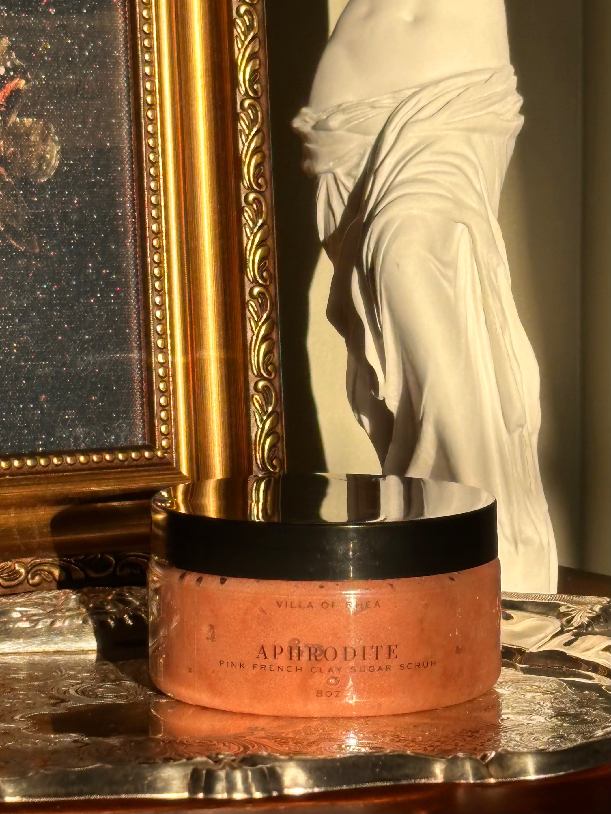Sugar scrub named 'Aphrodite' on a reflective surface with classical sculpture in the background .Vintage style. Greek Mythology Inspired. Luxury body scrub. handmade body scrubs.