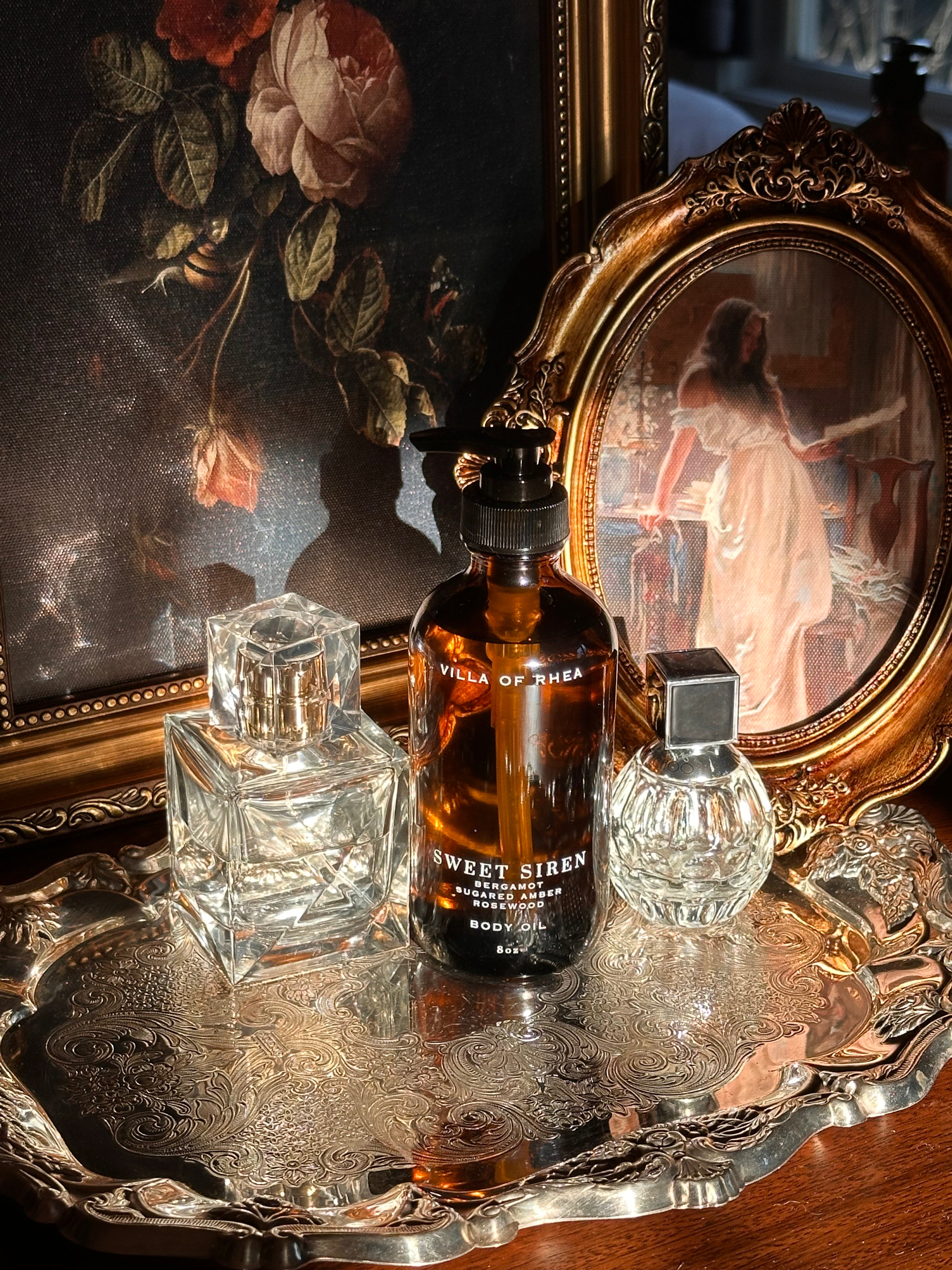 Villa of Rhea Sweet Siren body oil bottle on a reflective surface with decorative background.Sweet Siren Body Oil. Bergamot, Sandalwood, Amber, Pear scent. All Natural Skin Care. Non-Toxic Body Care, Greek Mythology Inspired. Chic Skincare. Chic Aesthetic. Minimalistic Aesthetic