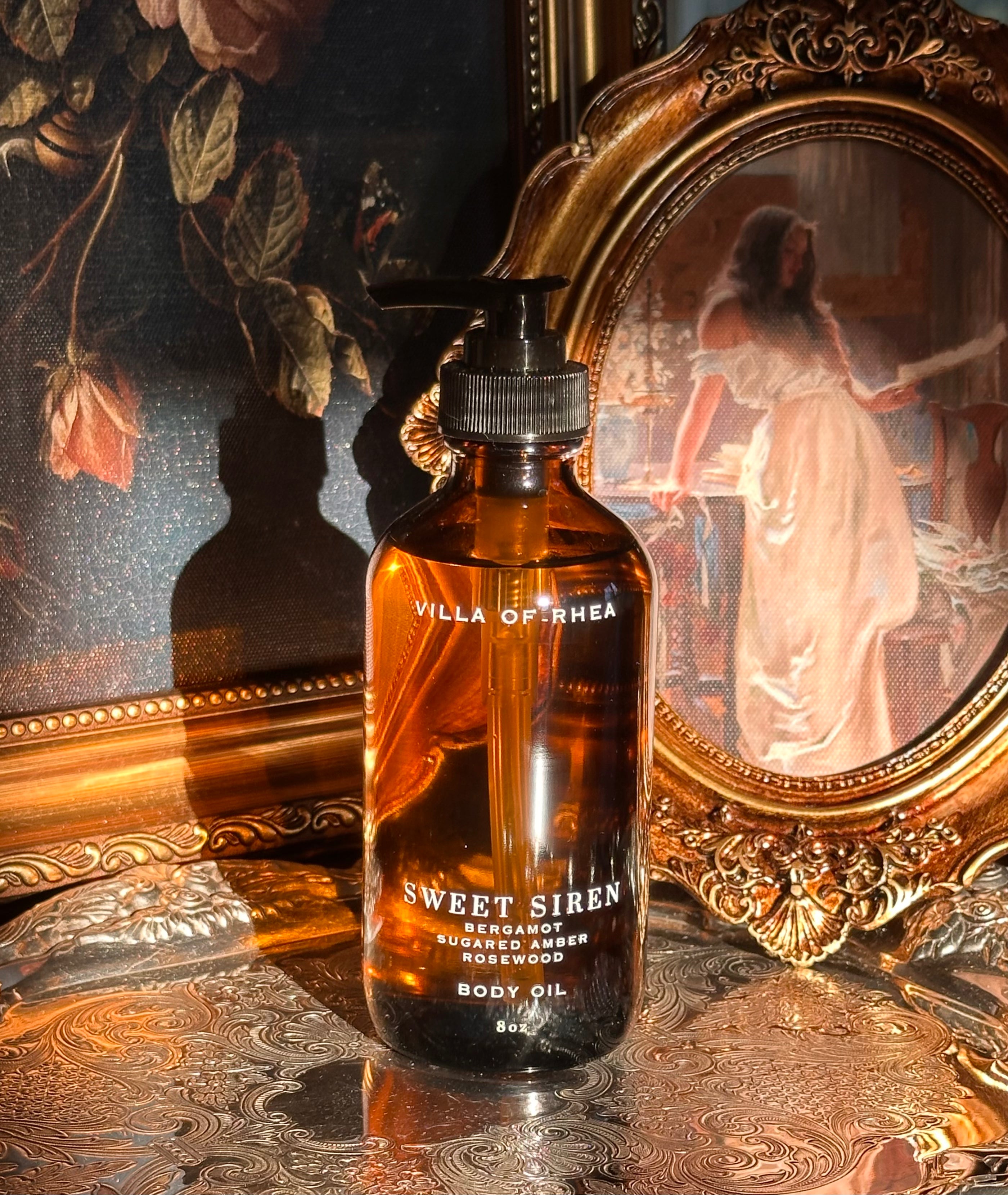 Villa of Rhea Sweet Siren body oil bottle on a reflective surface with decorative background.Sweet Siren Body Oil. Bergamot, Sandalwood, Amber, Pear scent. All Natural Skin Care. Non-Toxic Body Care, Greek Mythology Inspired. Chic Skincare. Chic Aesthetic. Minimalistic Aesthetic